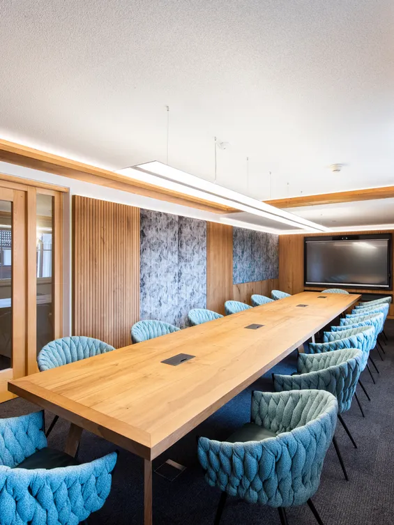 Moderner Konferenzraum mit langem Holztisch, blauen Stühlen und großem Bildschirm